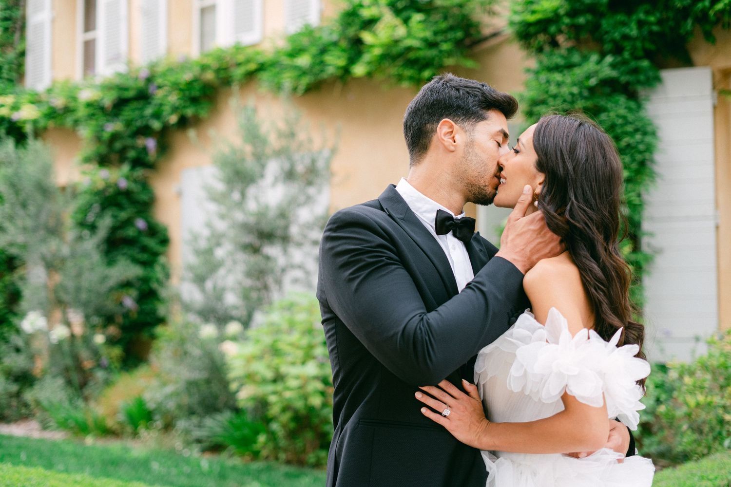 A couple shares a romantic kiss outdoors, groom in black tuxedo and bride in white off-shoulder gown before ivy-covered wall.