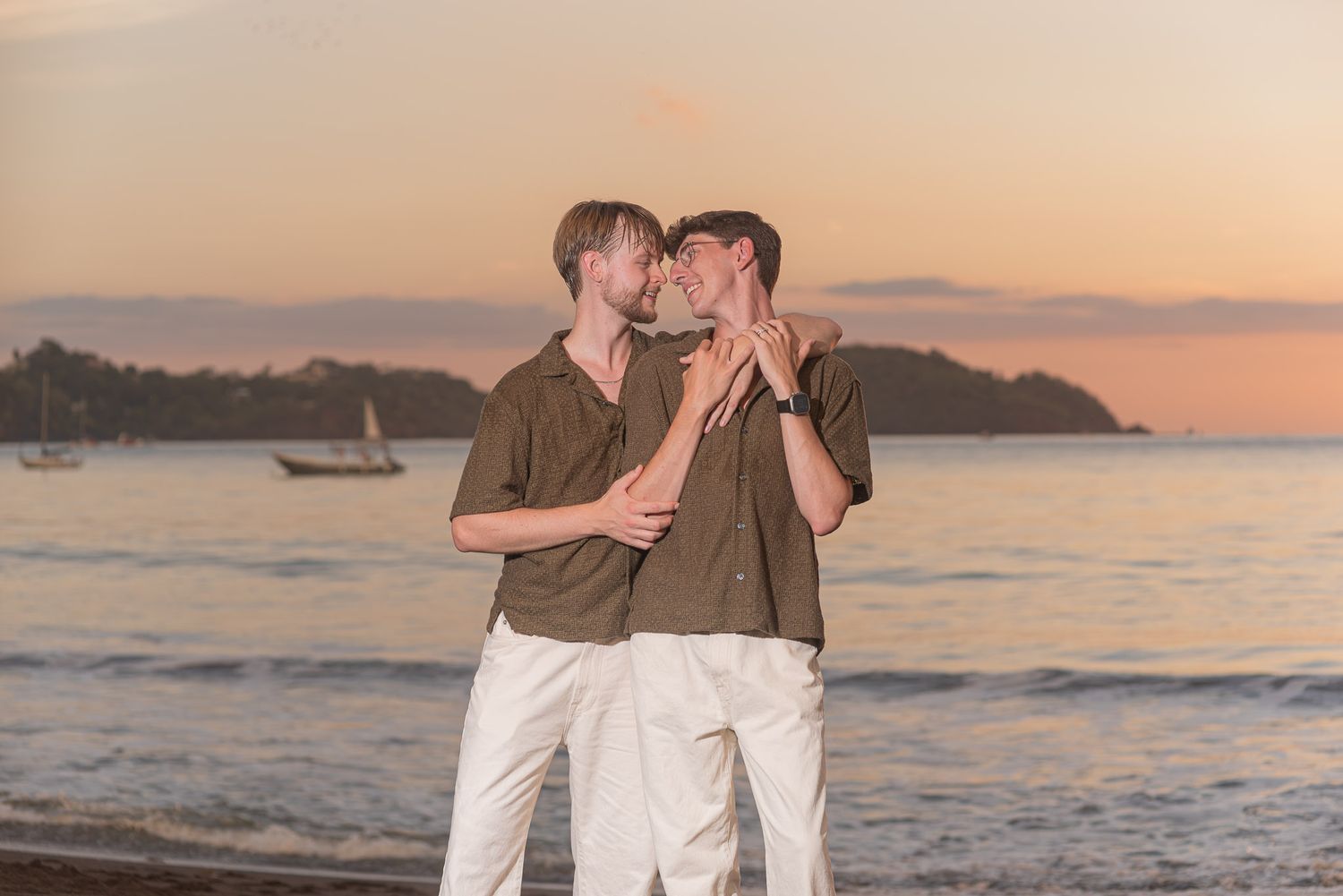 Two men sharing a romantic moment on a beach at sunset with sailboats and hills in the background.
