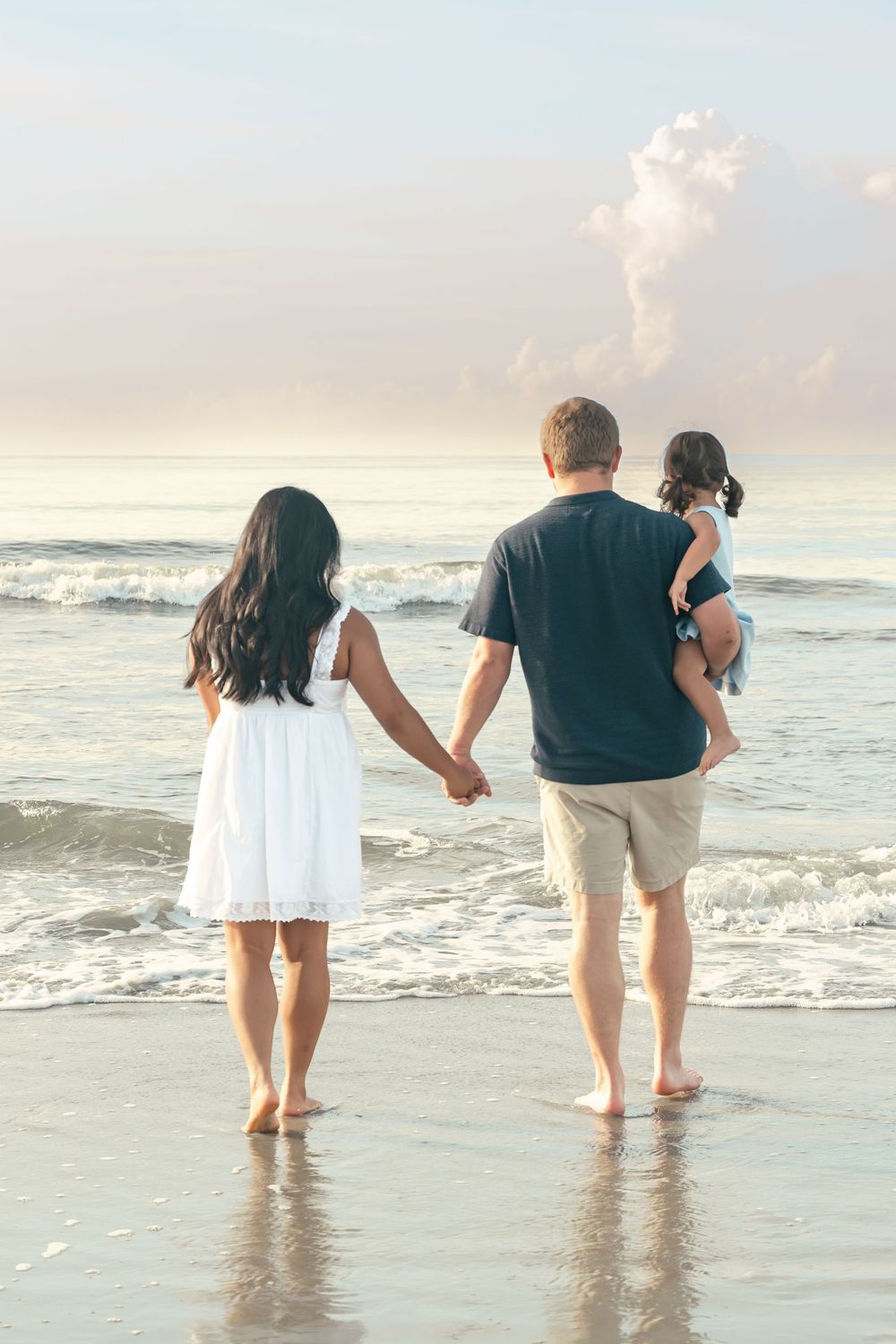 Family of 3 standing on the beach at sunrise, blue and pink sky over water with waves crashing at their feet