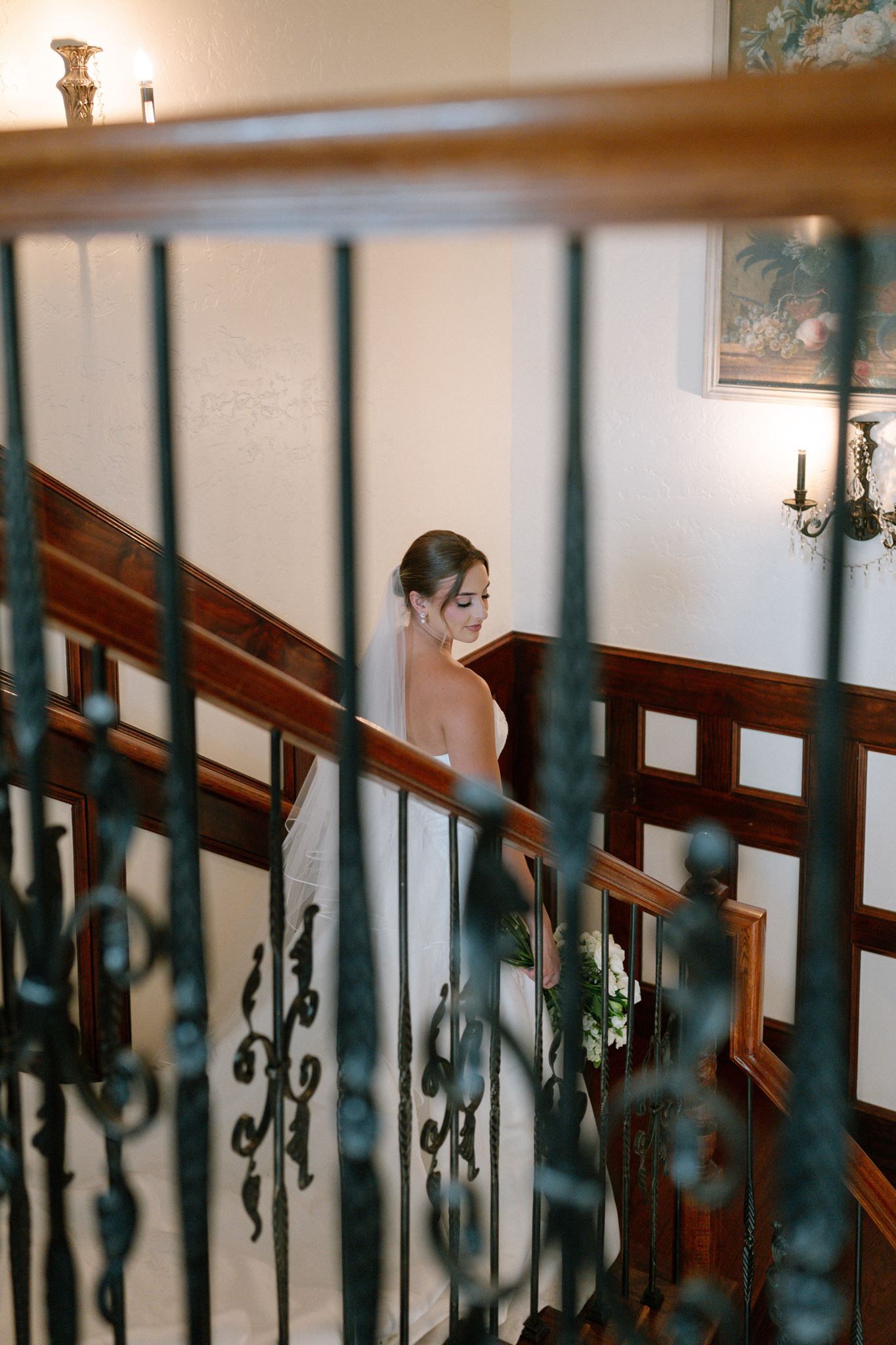 Bride in white gown descending elegant staircase with ornate iron railings and warm wood accents in a classic venue.