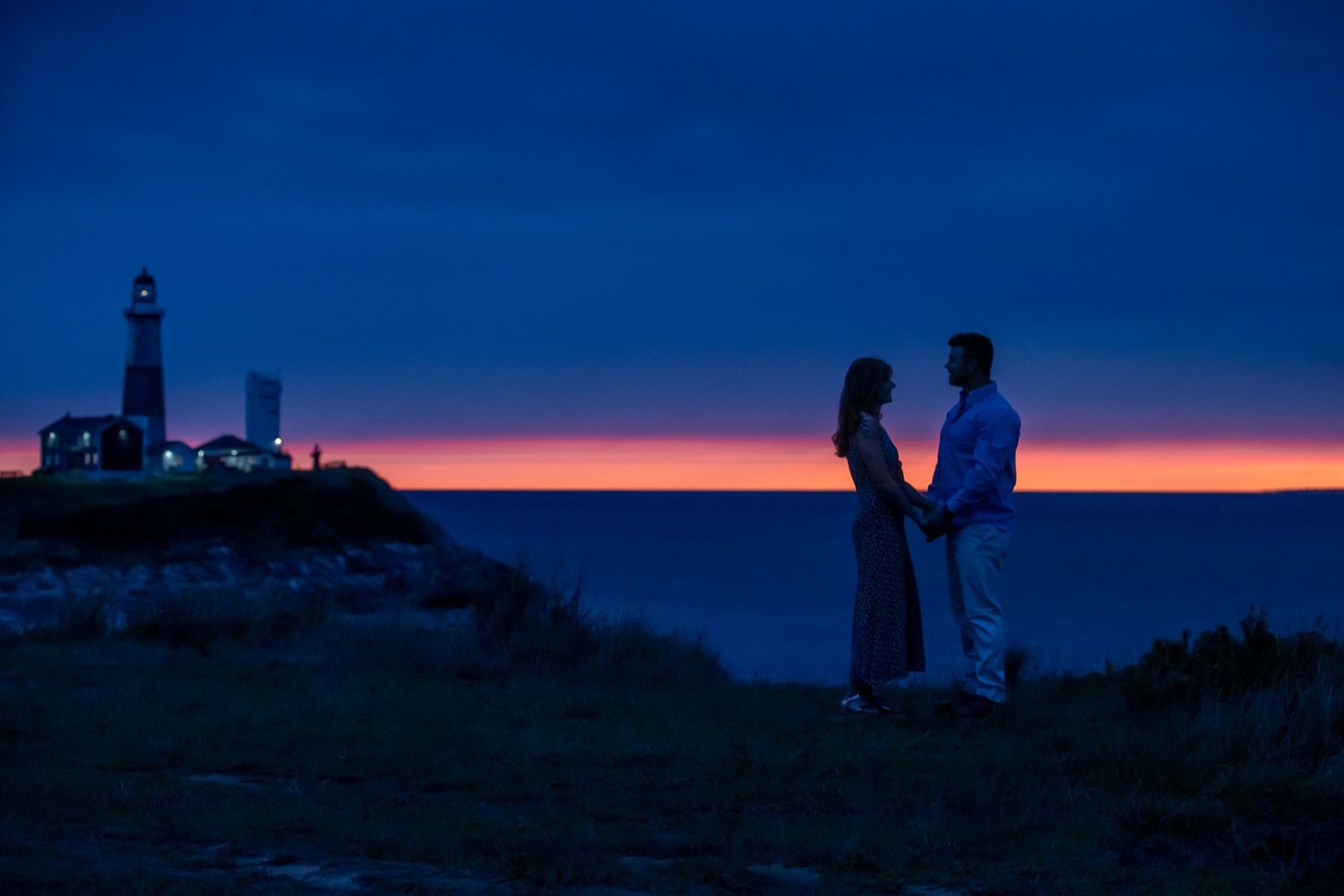 Couple holding hands at sunset near a lighthouse with vibrant orange horizon over the ocean.