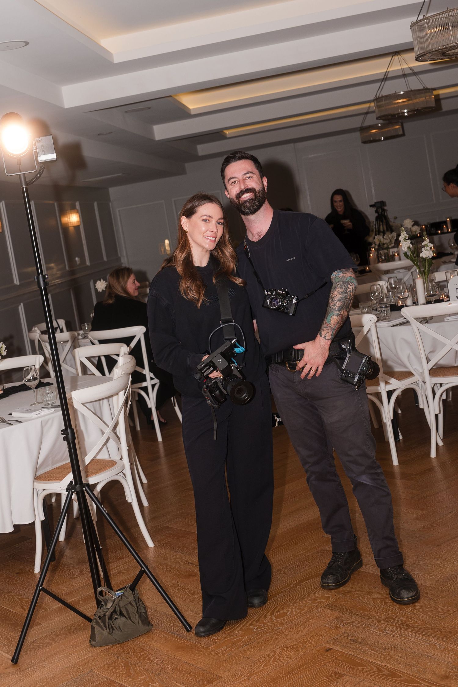 Two photographers in black attire pose with cameras at an elegant event venue with white chairs and dining tables.