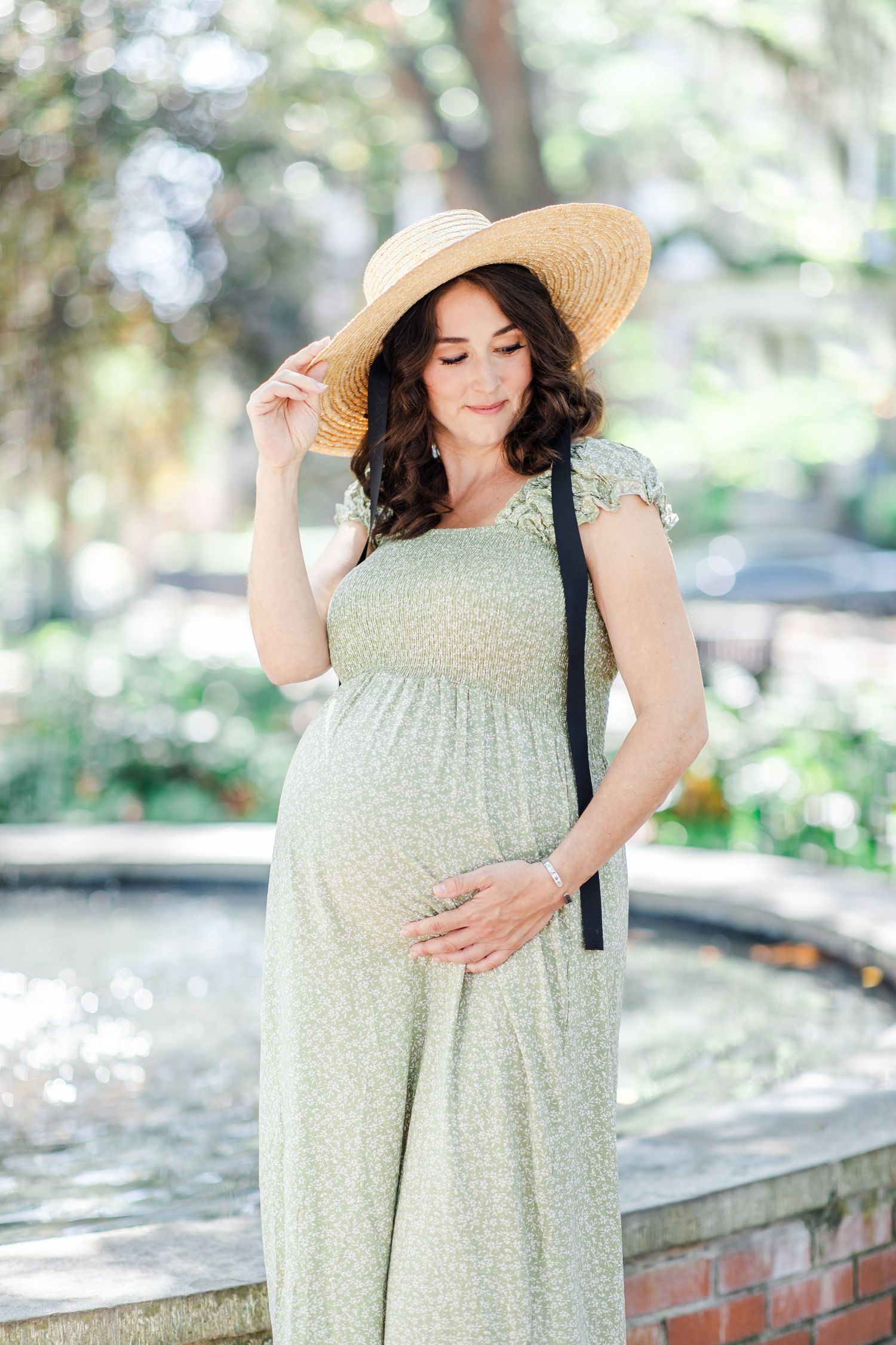 Pregnant woman in sage green floral dress and straw hat cradling baby bump outdoors near fountain.
