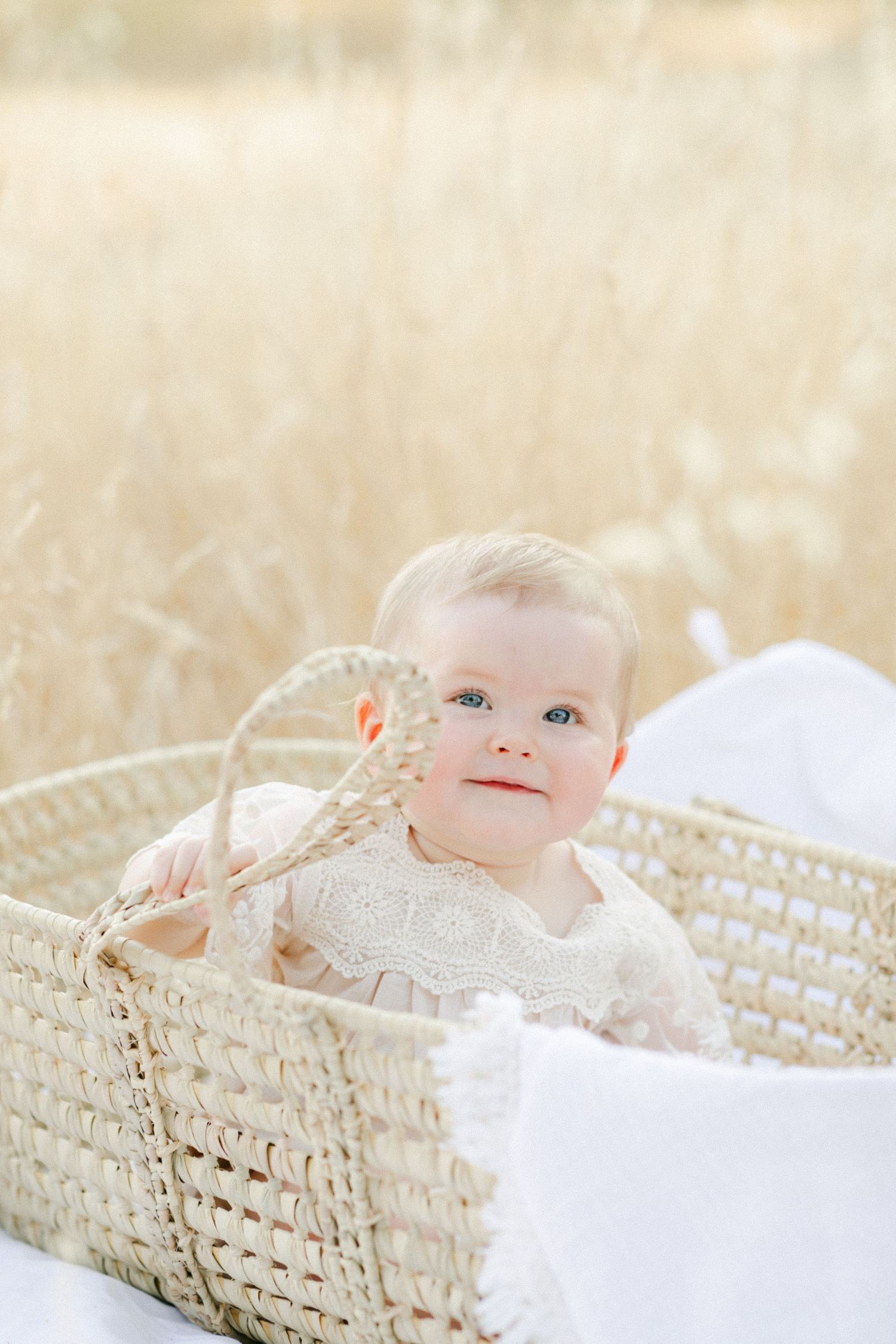 Smiling baby in a wicker Moses basket surrounded by golden wheat fields, wearing a cream lace dress.
