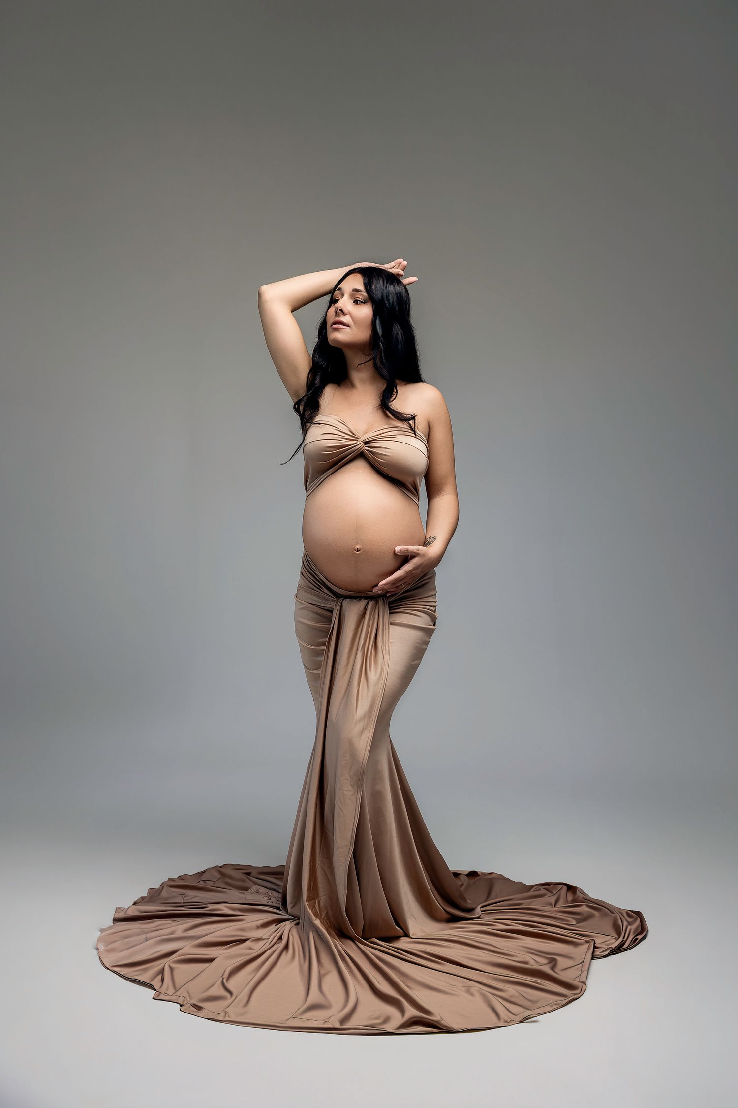 Pregnant woman in elegant tan mermaid gown posing gracefully in studio, cradling baby bump with one hand raised.
