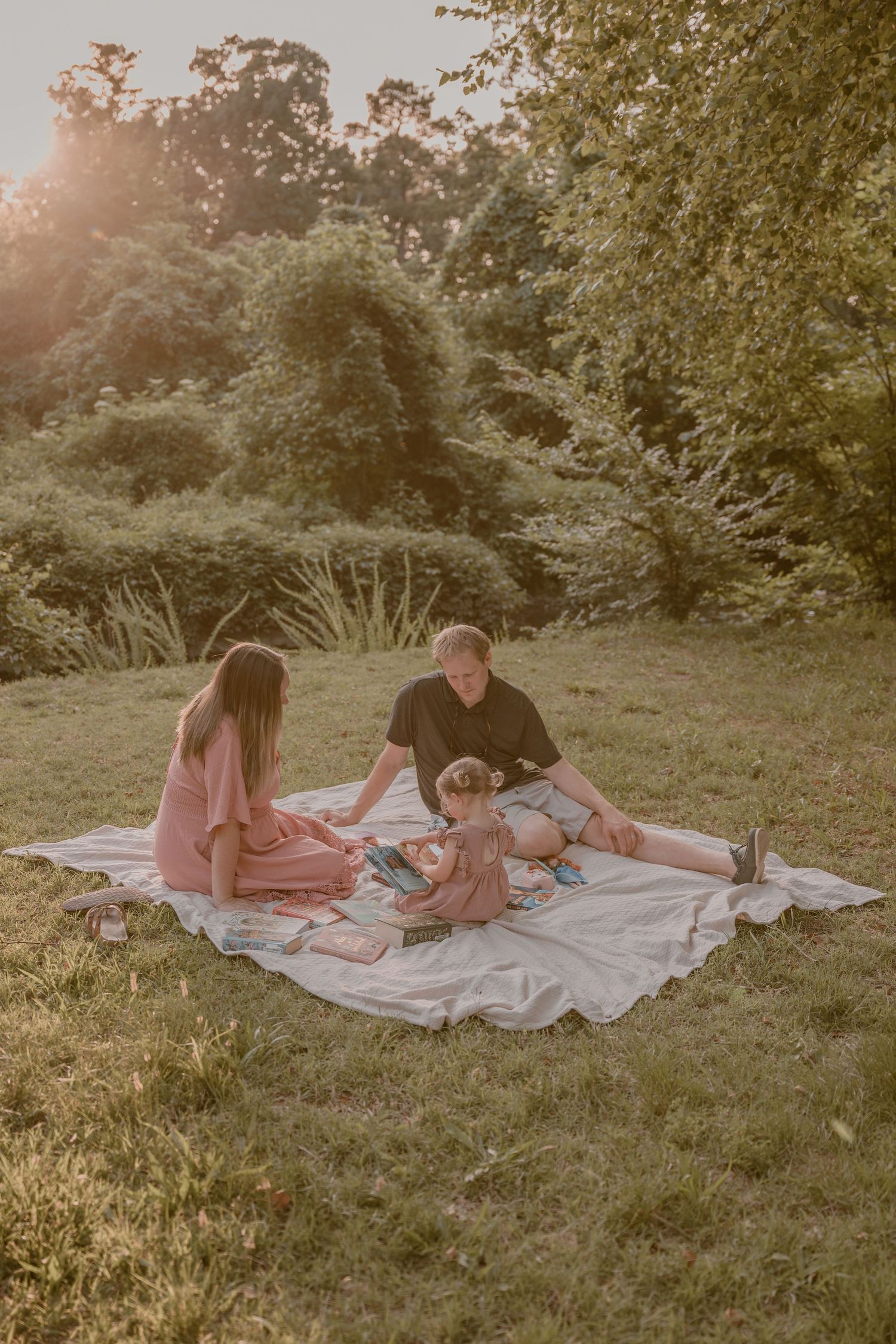 Family enjoying a sunny picnic on a blanket in a lush green park surrounded by trees.