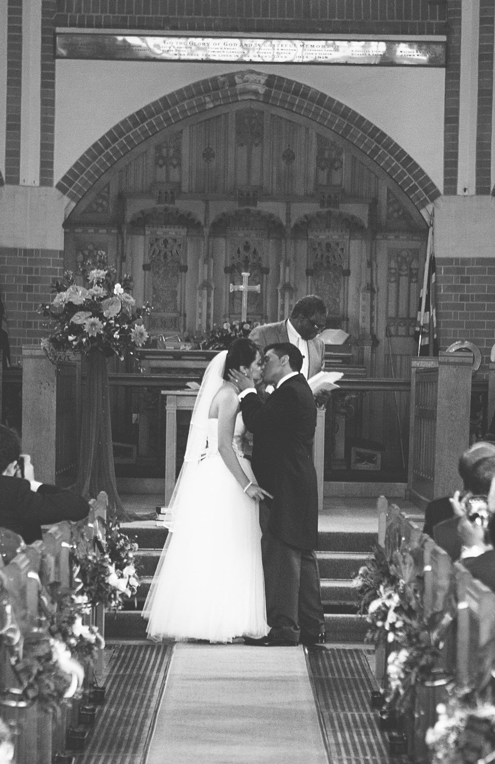 Black and white photo of a bride and groom sharing their first kiss at the altar during a church wedding ceremony.