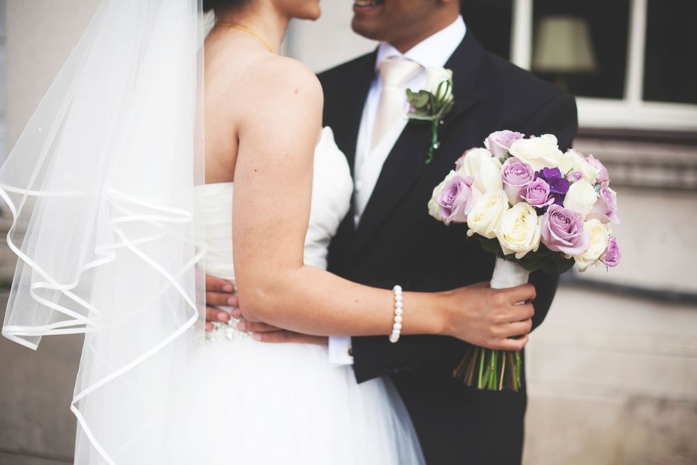 Bride and groom embracing, holding a bouquet of purple and white roses on their wedding day.