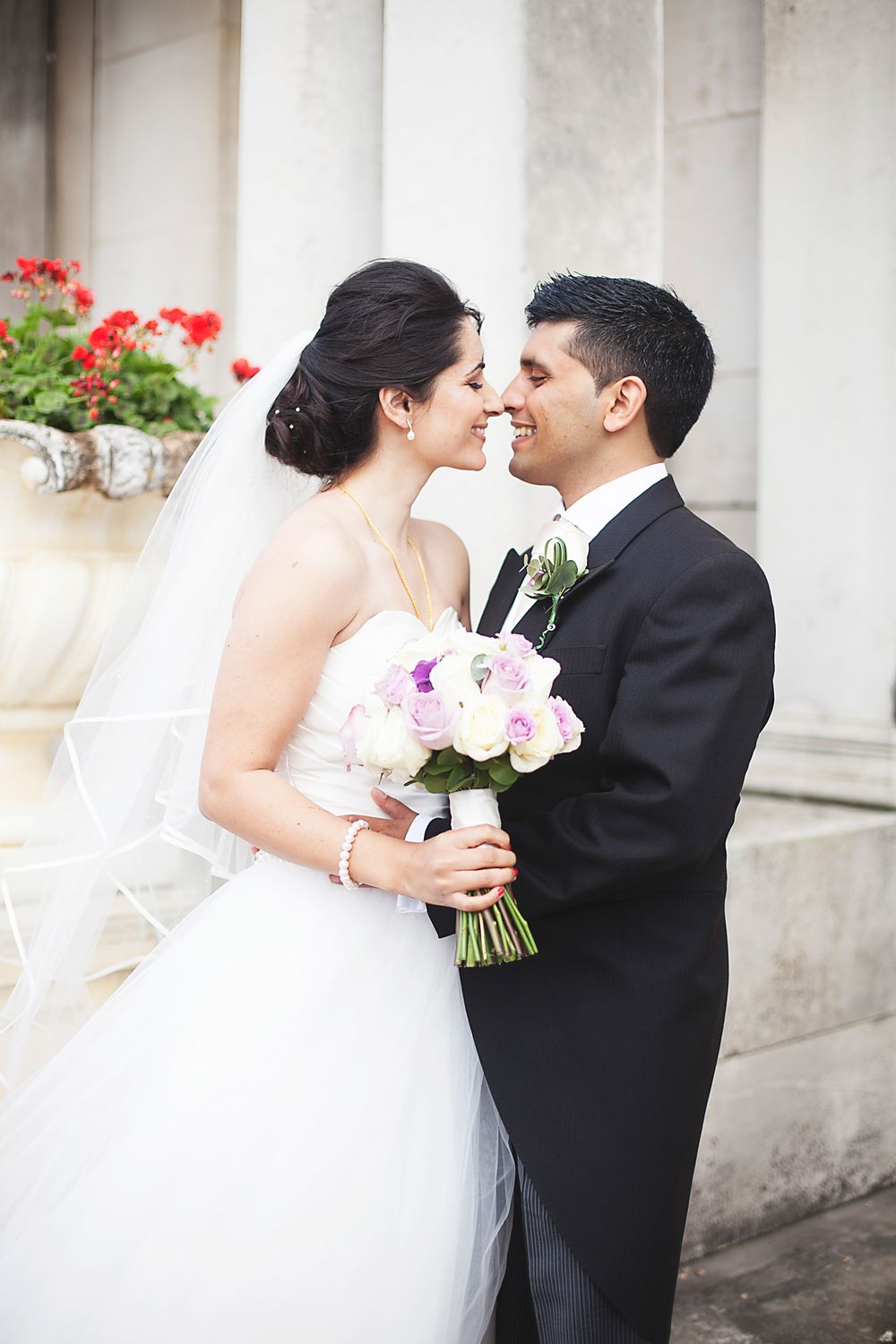 Bride and groom share a romantic moment, holding a pink and white bouquet, with red flowers and columns in the background.