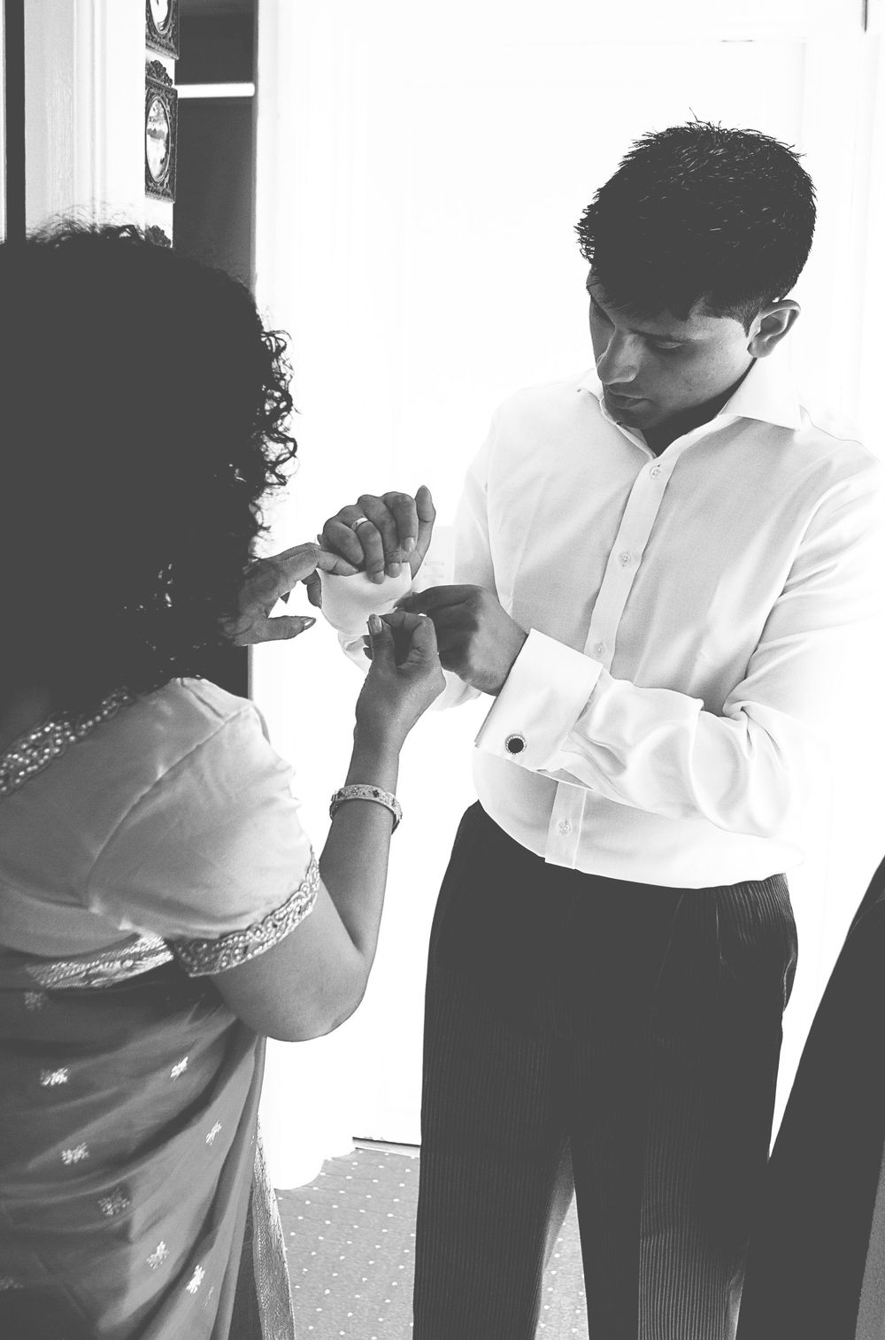 Black and white photo of a woman in a sari helping a man in a white dress shirt fasten his cufflinks.
