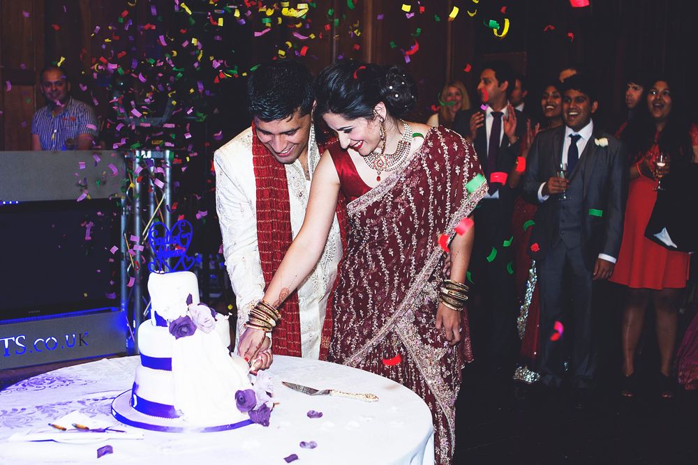 Indian couple in traditional attire cutting wedding cake at colorful confetti-filled reception celebration.