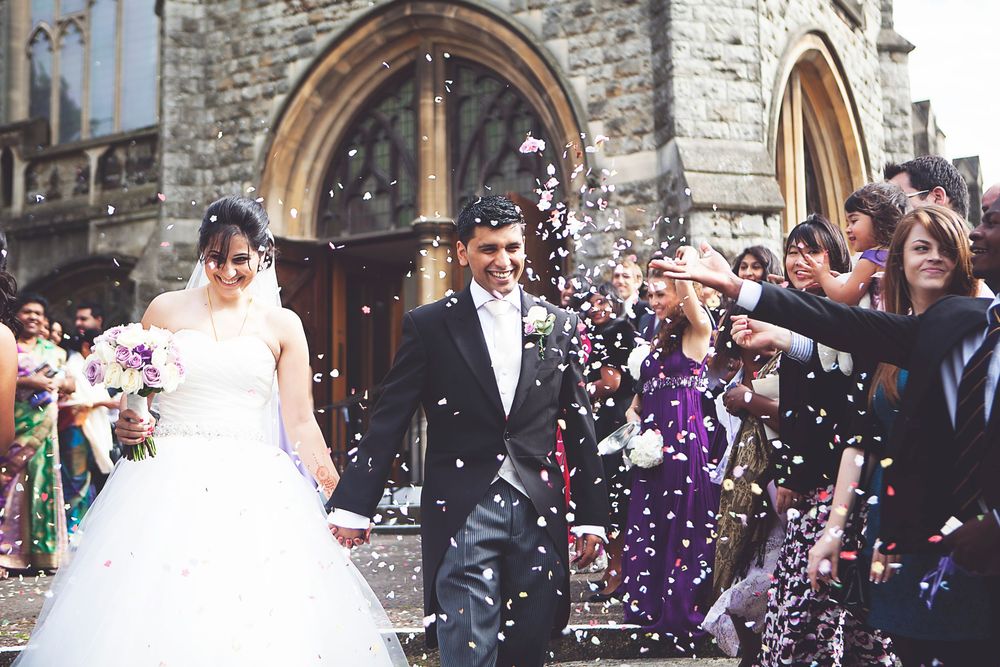 Bride and groom exit church as guests throw confetti during outdoor wedding ceremony.