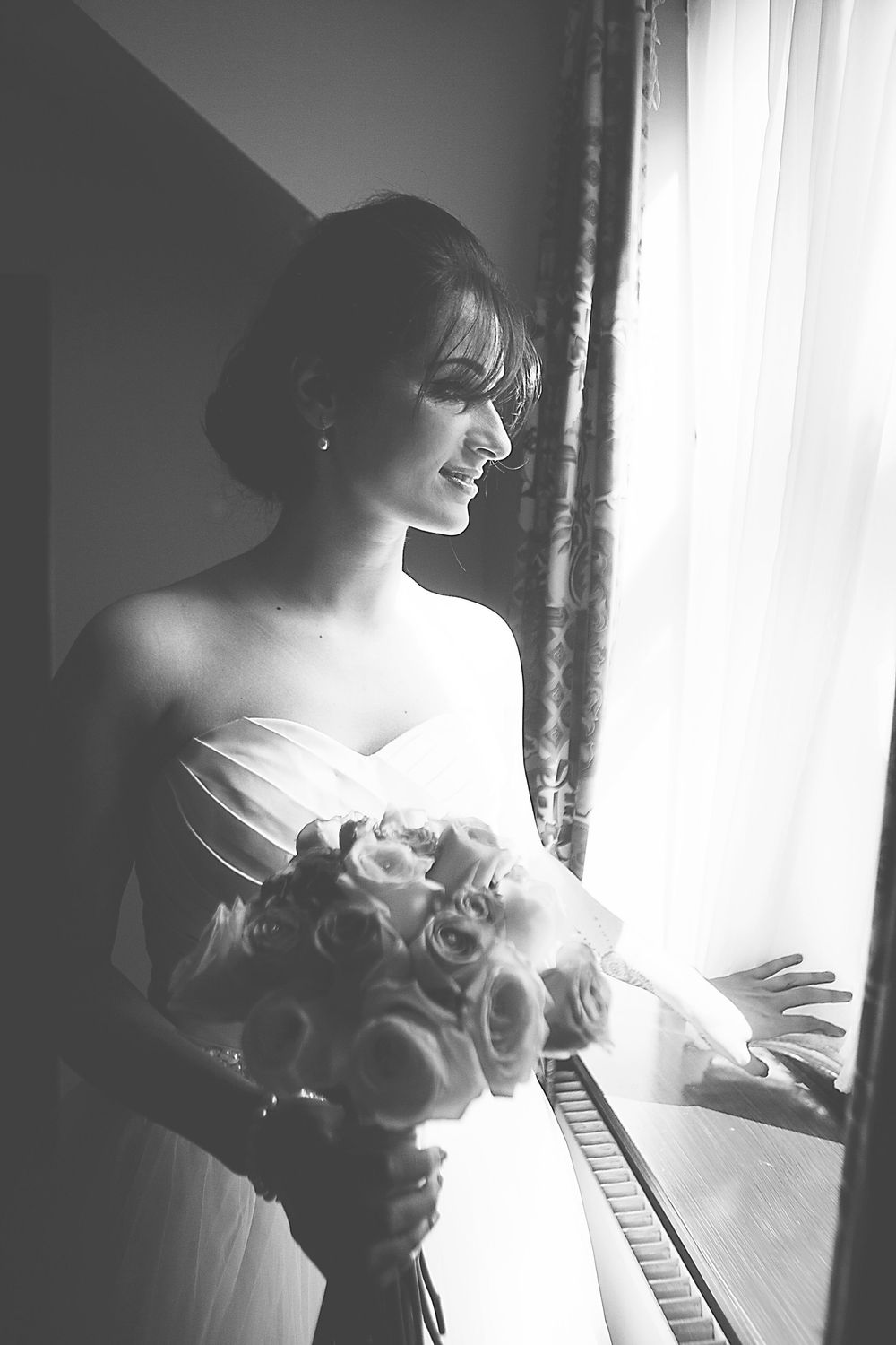Black and white photo of a bride in strapless gown holding rose bouquet, gazing out a sunlit window.