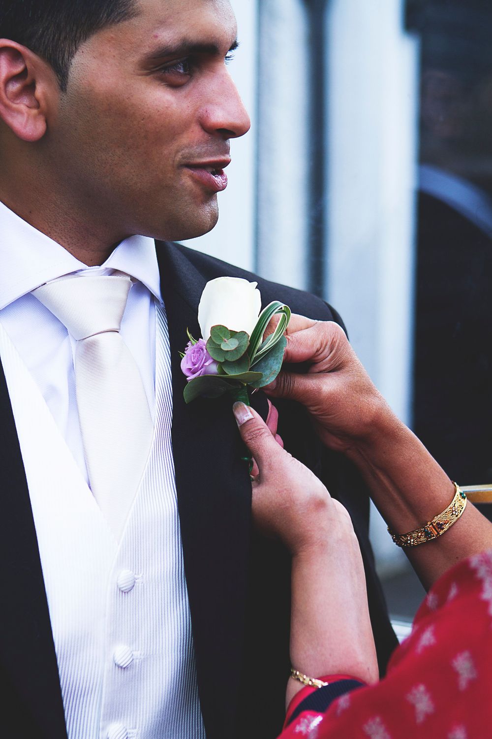A woman pins a white and purple boutonniere onto a groom's black suit lapel at a wedding ceremony.