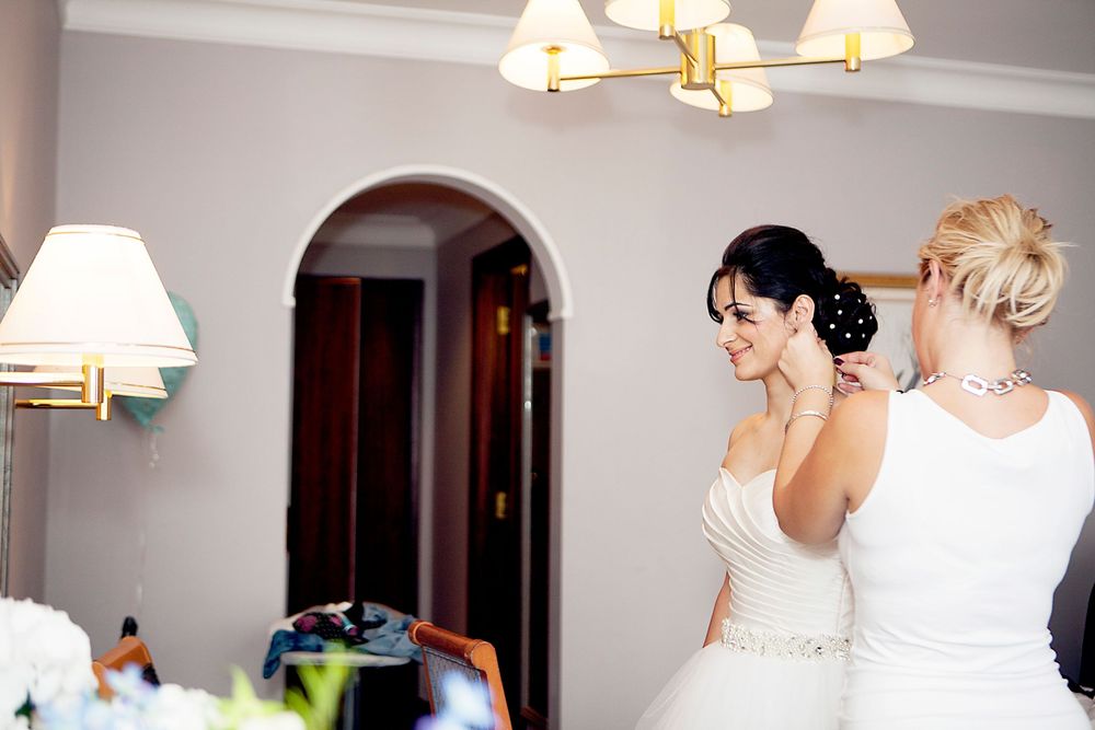 Two brides in white wedding gowns help each other prepare, sharing a tender moment in an elegant room.