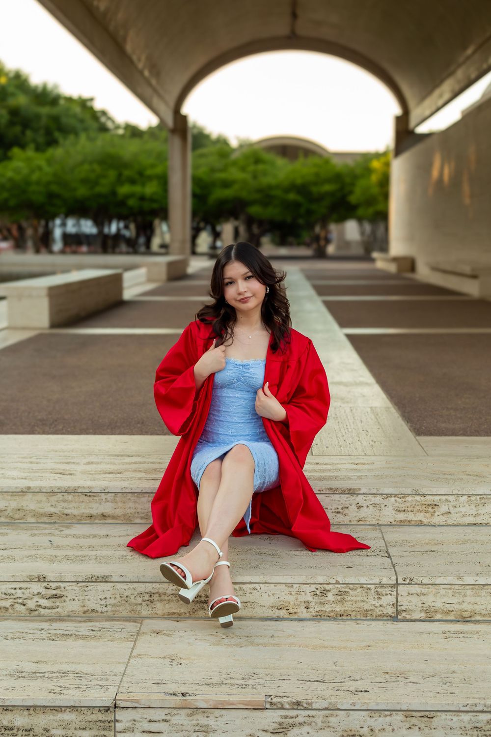 Young woman in red graduation gown and blue dress sitting on steps under arched structure with green trees.