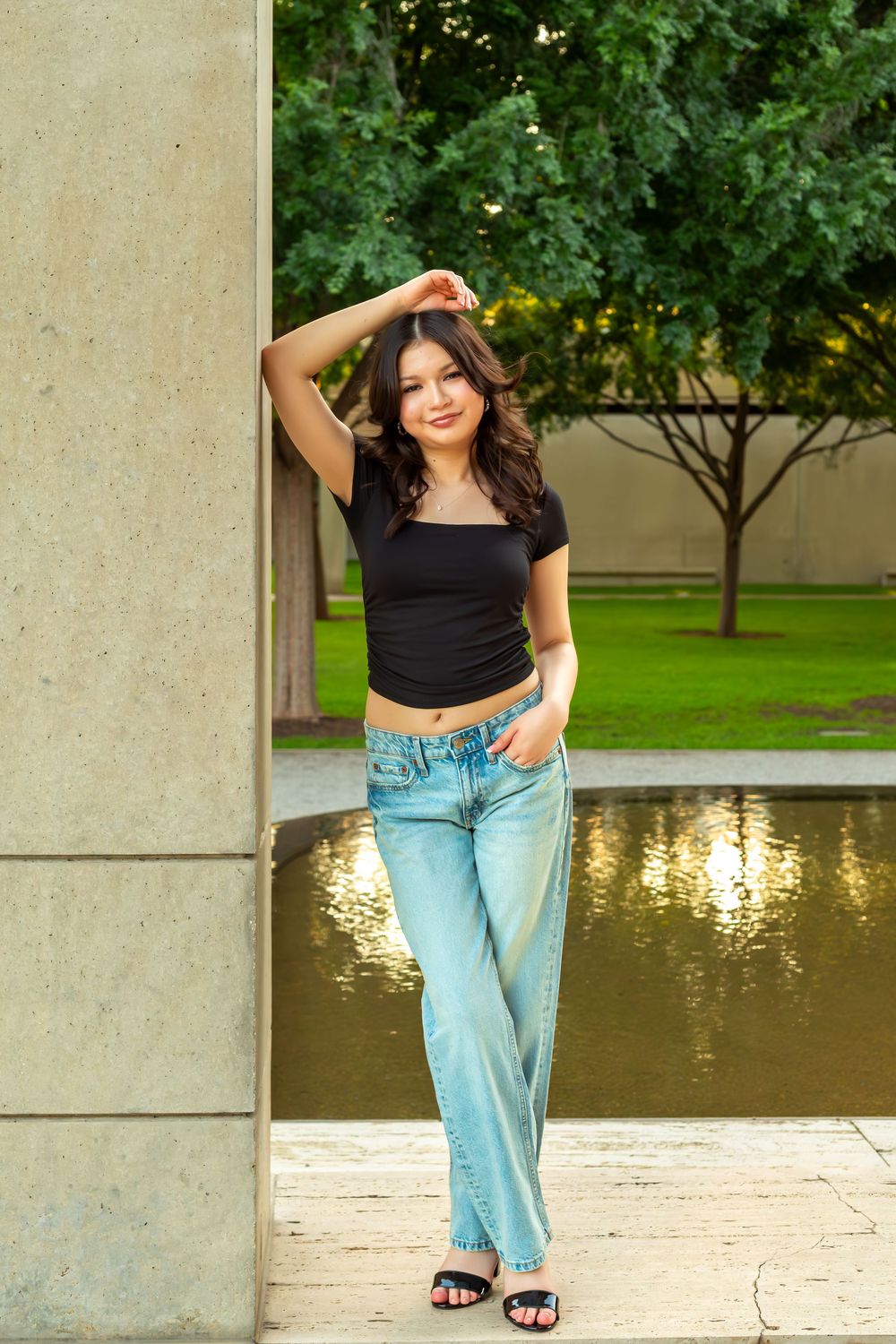 Young woman in black crop top and blue jeans posing by stone column near reflective pool outdoors.