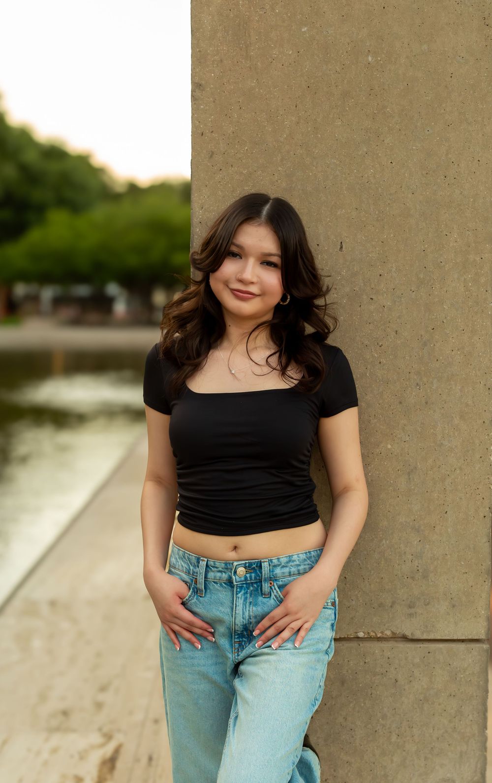 Young woman in black crop top and jeans leaning against stone column near water, smiling softly.