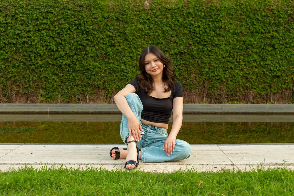 Young woman in black crop top and blue jeans sitting cross-legged on pavement beside green hedge outdoors.