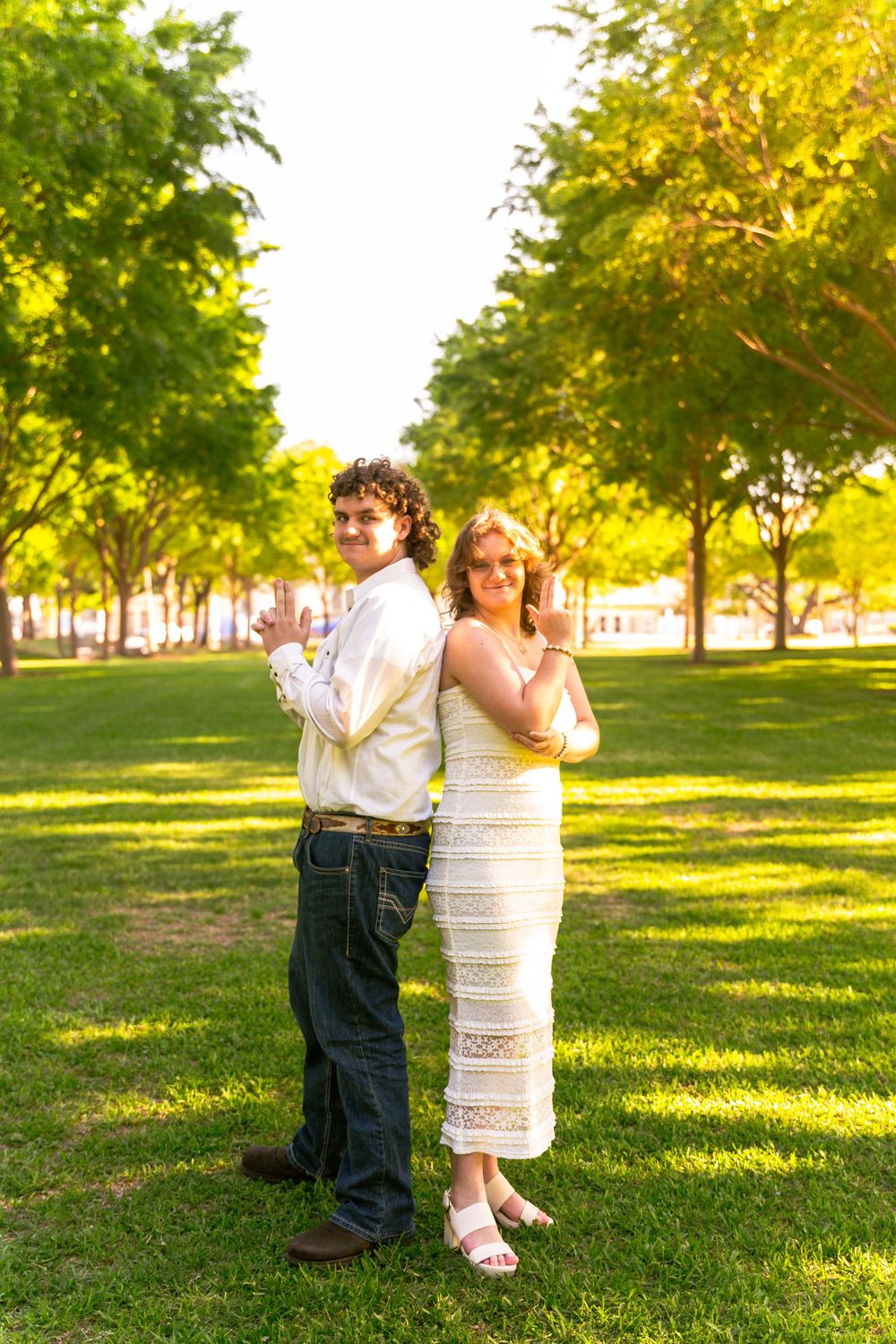 A couple stands back-to-back in a sunny park, the woman in a white dress and the man in jeans and white shirt.