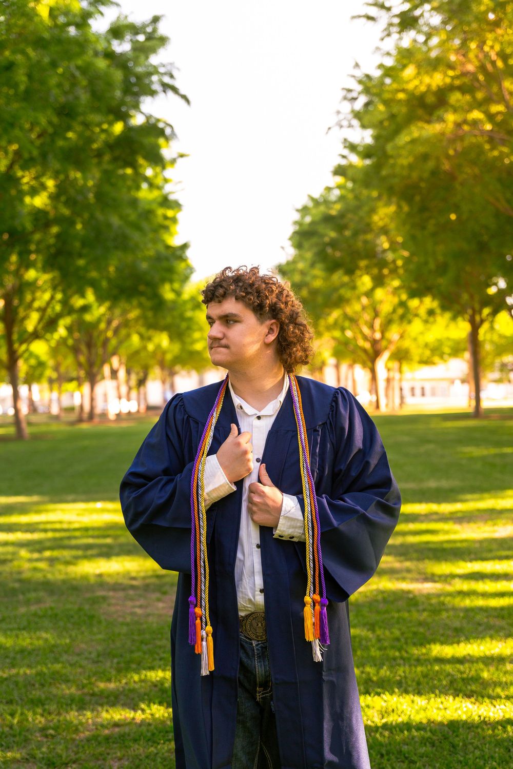 Young man in navy blue graduation gown with colorful honor cords poses outdoors in a tree-lined park.