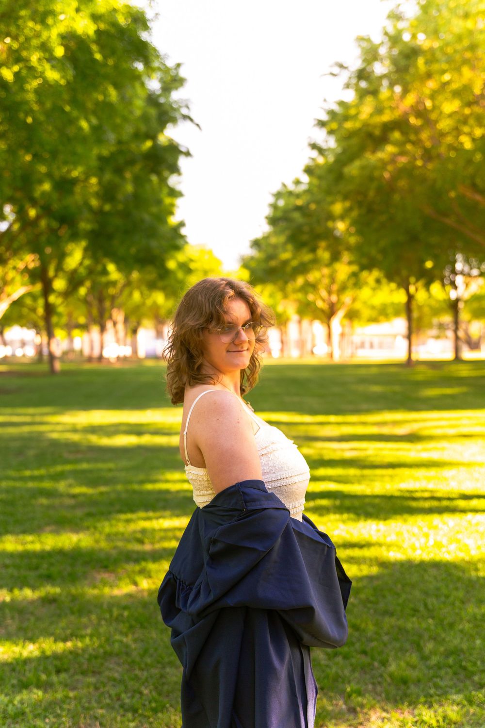 Young woman in white top and navy skirt smiling in a sunny green park with tree-lined path.