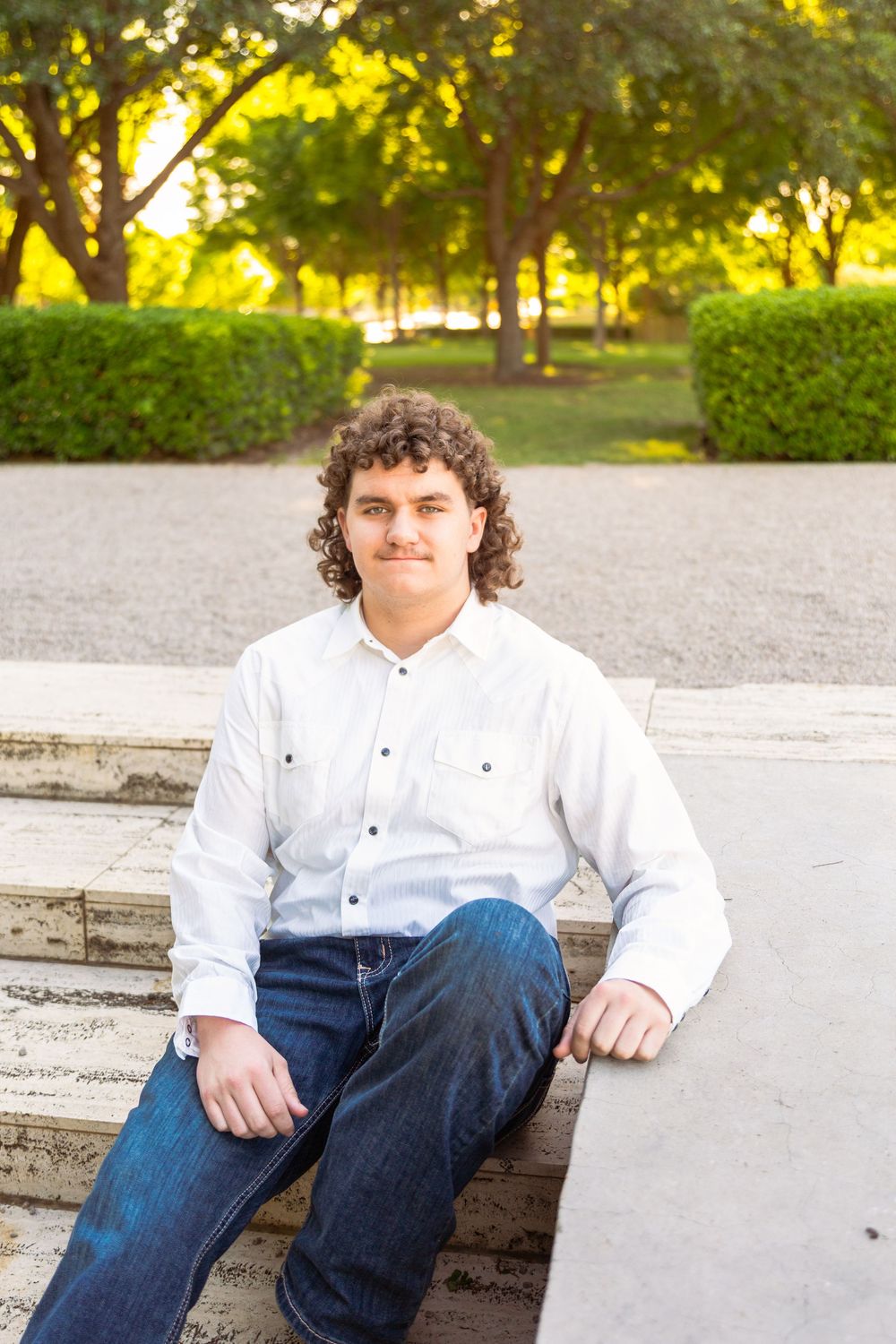 Young man with curly hair in white shirt and jeans sitting on stone steps outdoors with green trees behind him.