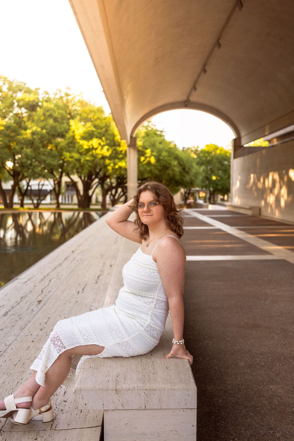 Young woman in white lace dress sitting on stone ledge under arched colonnade with golden sunlight and green trees.