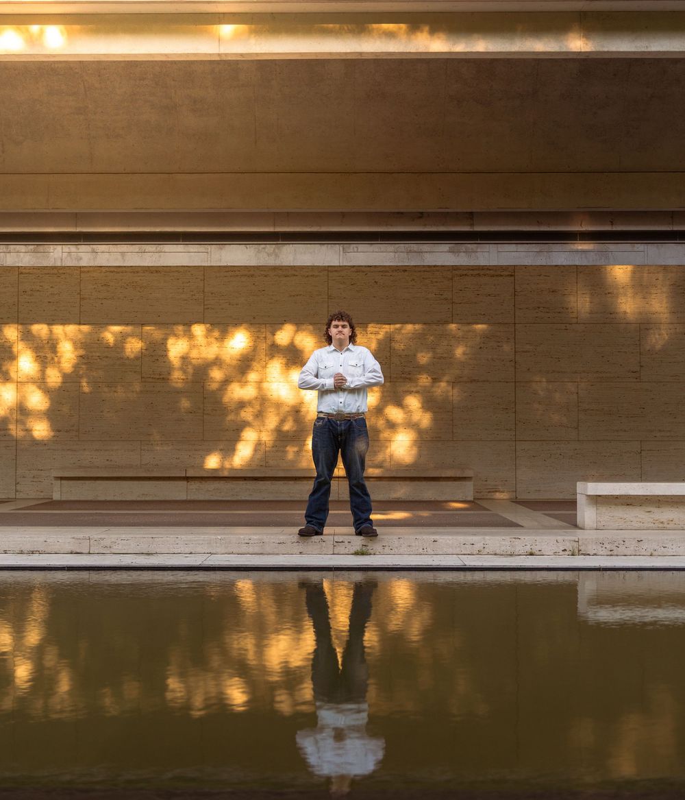 Man standing by reflective pool in modern architectural space with warm golden light and tree shadows.