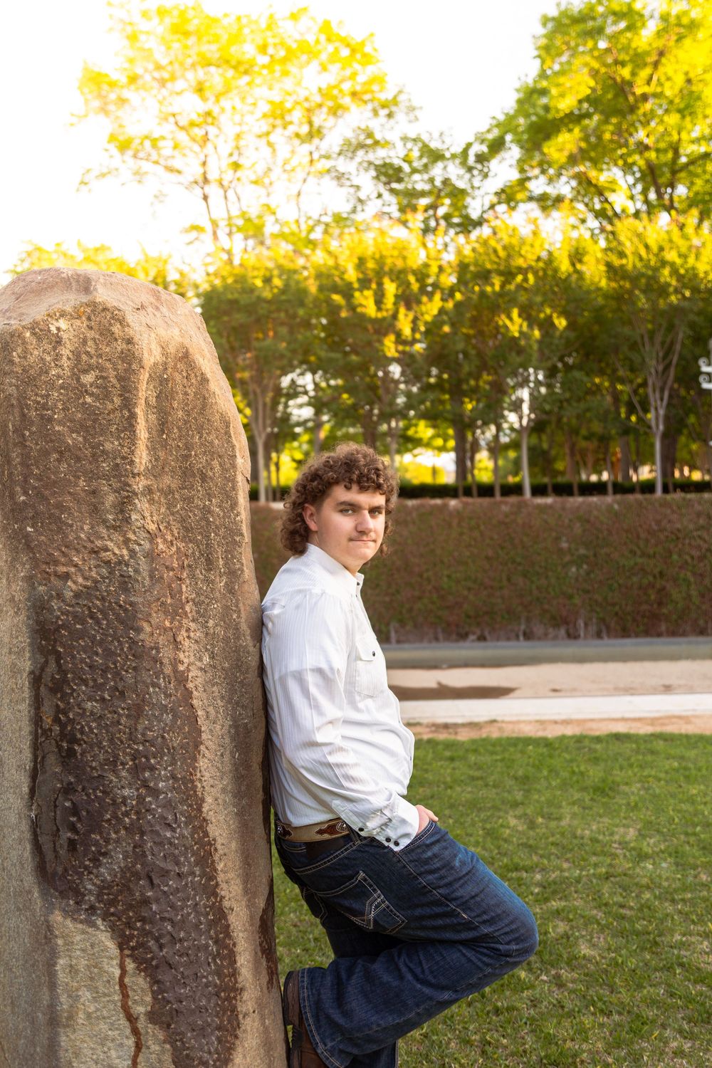 Young man in white shirt and jeans leaning against a large rock in a sunlit park setting.