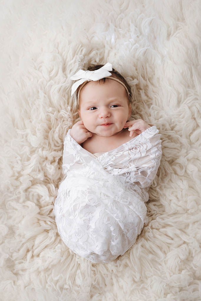 Newborn baby girl swaddled in white lace wrap with bow headband, lying on fluffy cream fur rug.