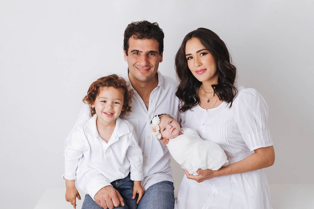 Family portrait with parents and two young children dressed in white against a light background.