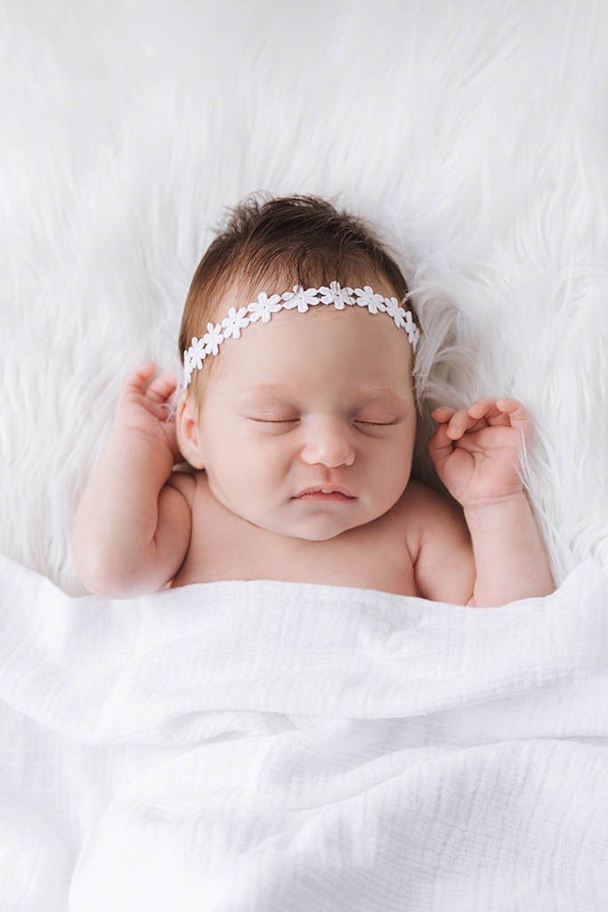 Sleeping newborn baby girl wearing a white floral headband, wrapped in a soft white blanket on a fluffy white fur background.