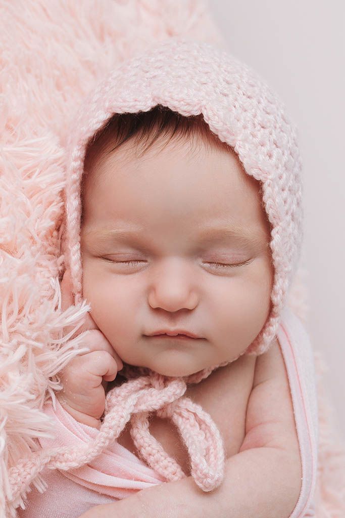 Sleeping newborn baby girl wearing a pink knit bonnet, wrapped in a soft pink fluffy blanket.