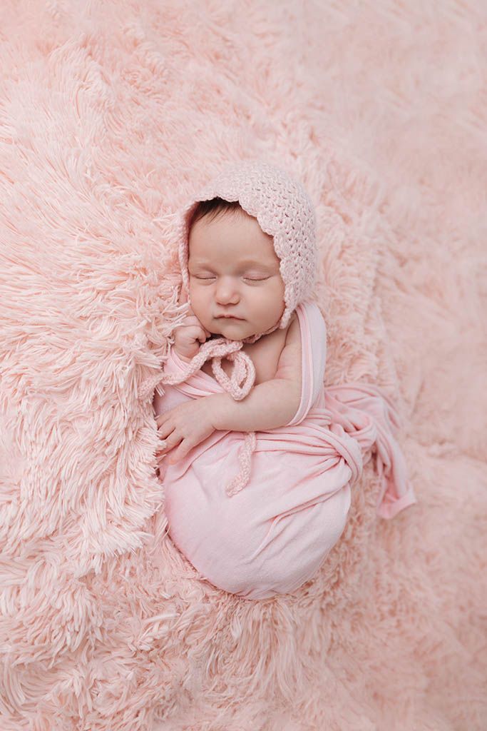 Sleeping newborn baby girl wrapped in pink swaddle with knit bonnet on a soft pink fluffy blanket.