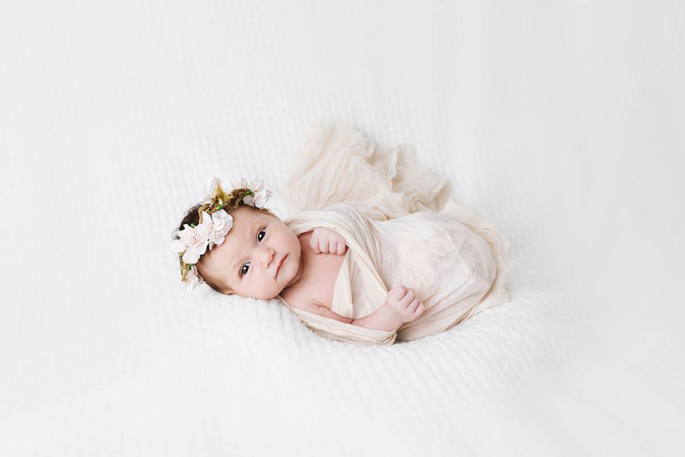 Newborn baby girl wrapped in cream fabric with floral crown lying on white blanket.