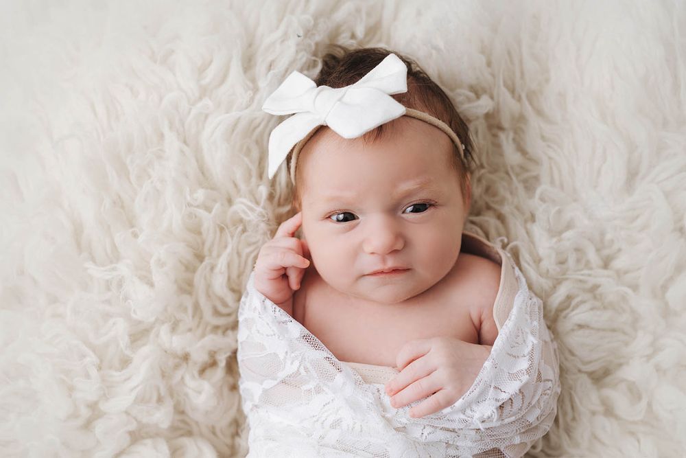Newborn baby girl wrapped in white lace swaddle with bow headband lying on fluffy cream fur backdrop.