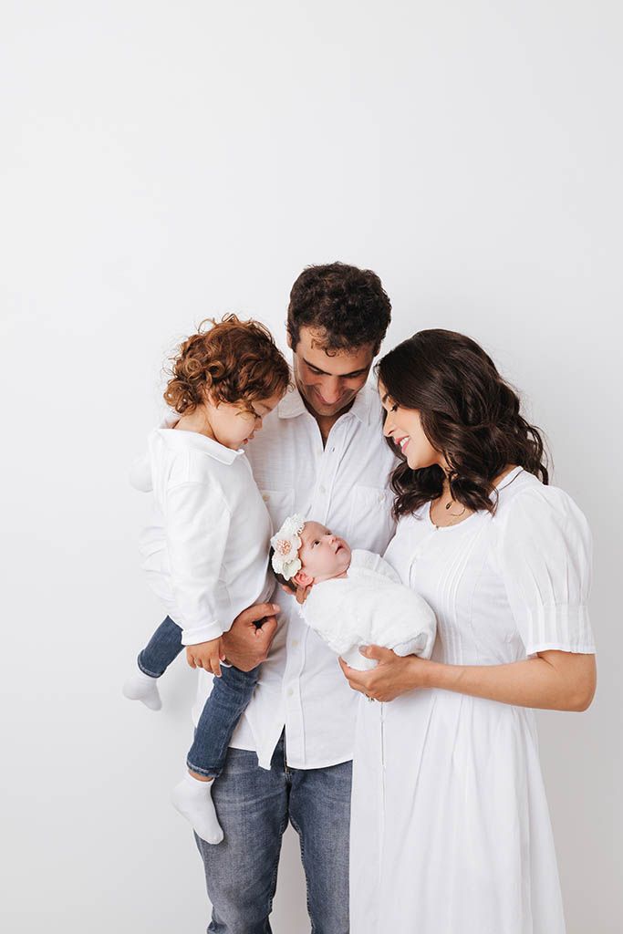 Family portrait with parents holding newborn baby and toddler in white outfits.