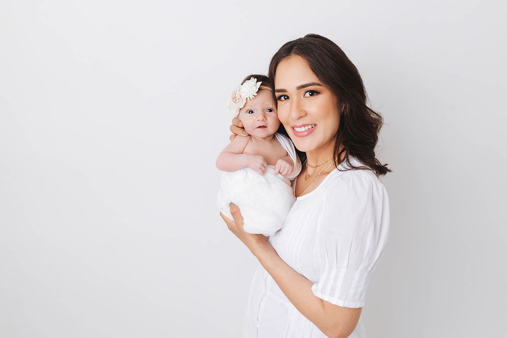 Smiling brunette mother in white dress holding her newborn baby girl.