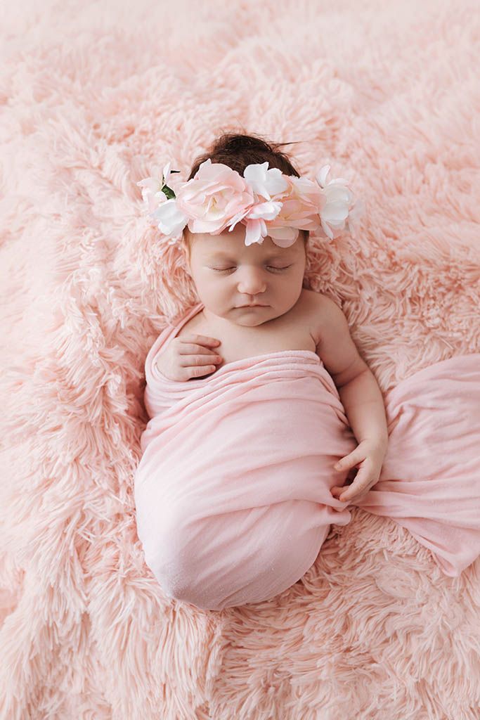 Sleeping newborn baby girl swaddled in pink wrap with flower headband on fluffy pink rug.