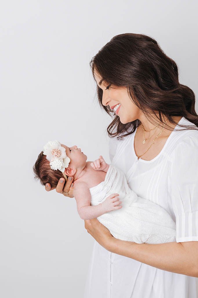 Brunette mother in white dress lovingly gazing at her newborn baby with floral headband.