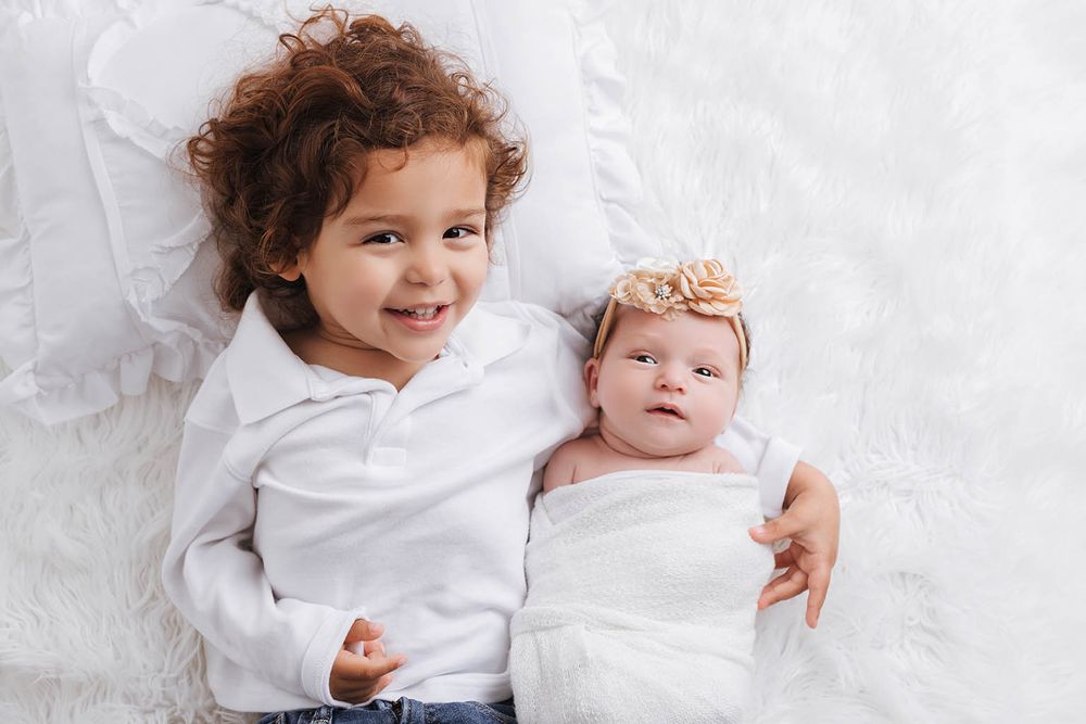 Smiling toddler with curly hair lying beside a newborn baby with a floral headband.