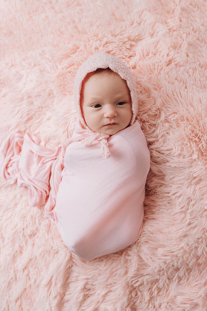 Newborn baby swaddled in pink wrap with knit bonnet on fluffy pink blanket.