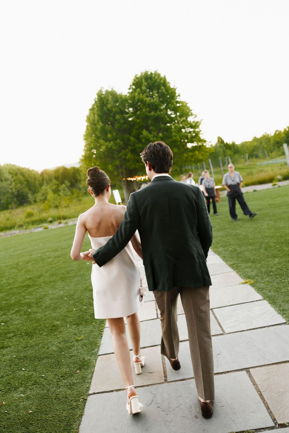 Bride and groom walking hand in hand down a stone path through lush green wedding grounds.