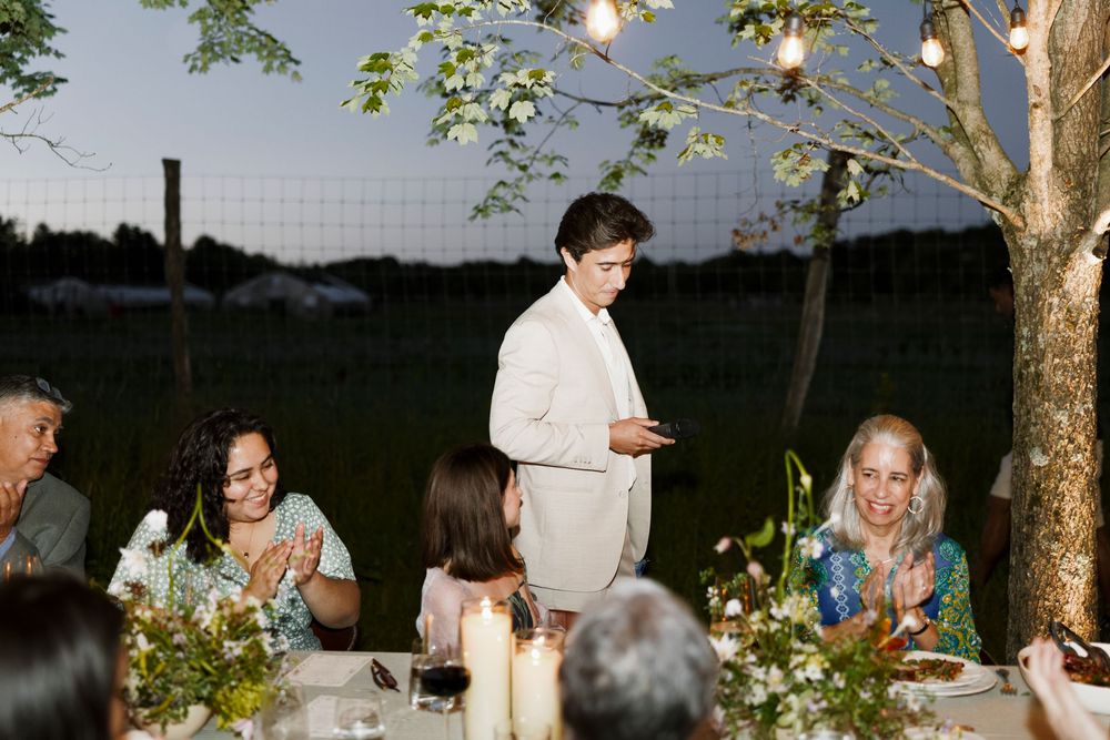 Person in white suit giving speech at outdoor wedding reception table at dusk.