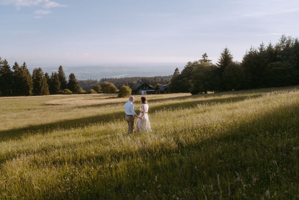 Couple walks hand in hand through a lush green meadow with trees and ocean view in the background.