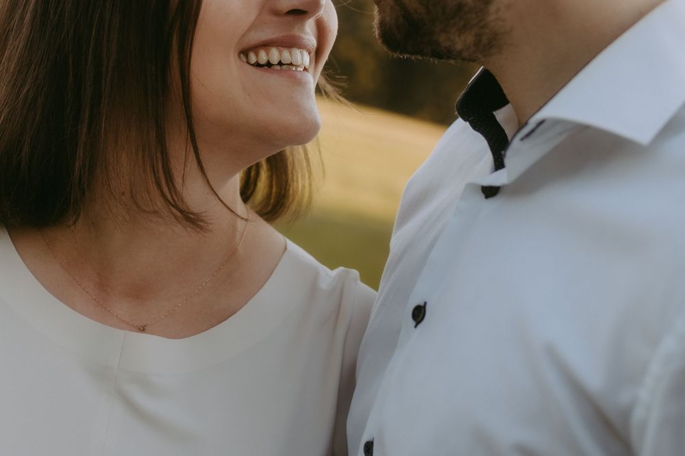 Couple smiling and facing each other outdoors, woman in white top and man in white dress shirt with bow tie.