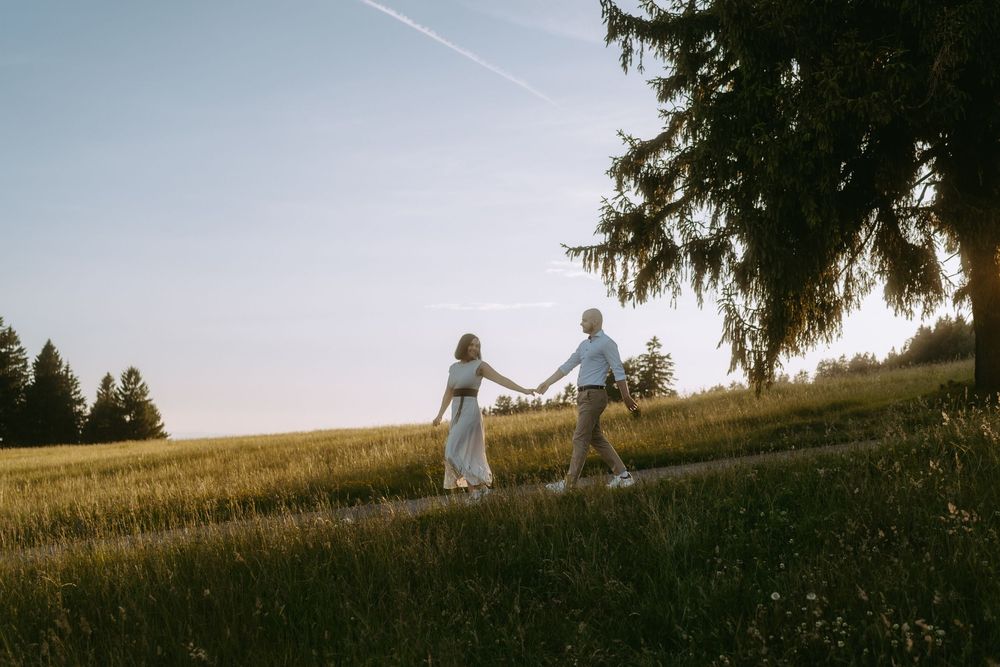 Couple holding hands walking through a sunlit green meadow at golden hour with tall trees in the background.