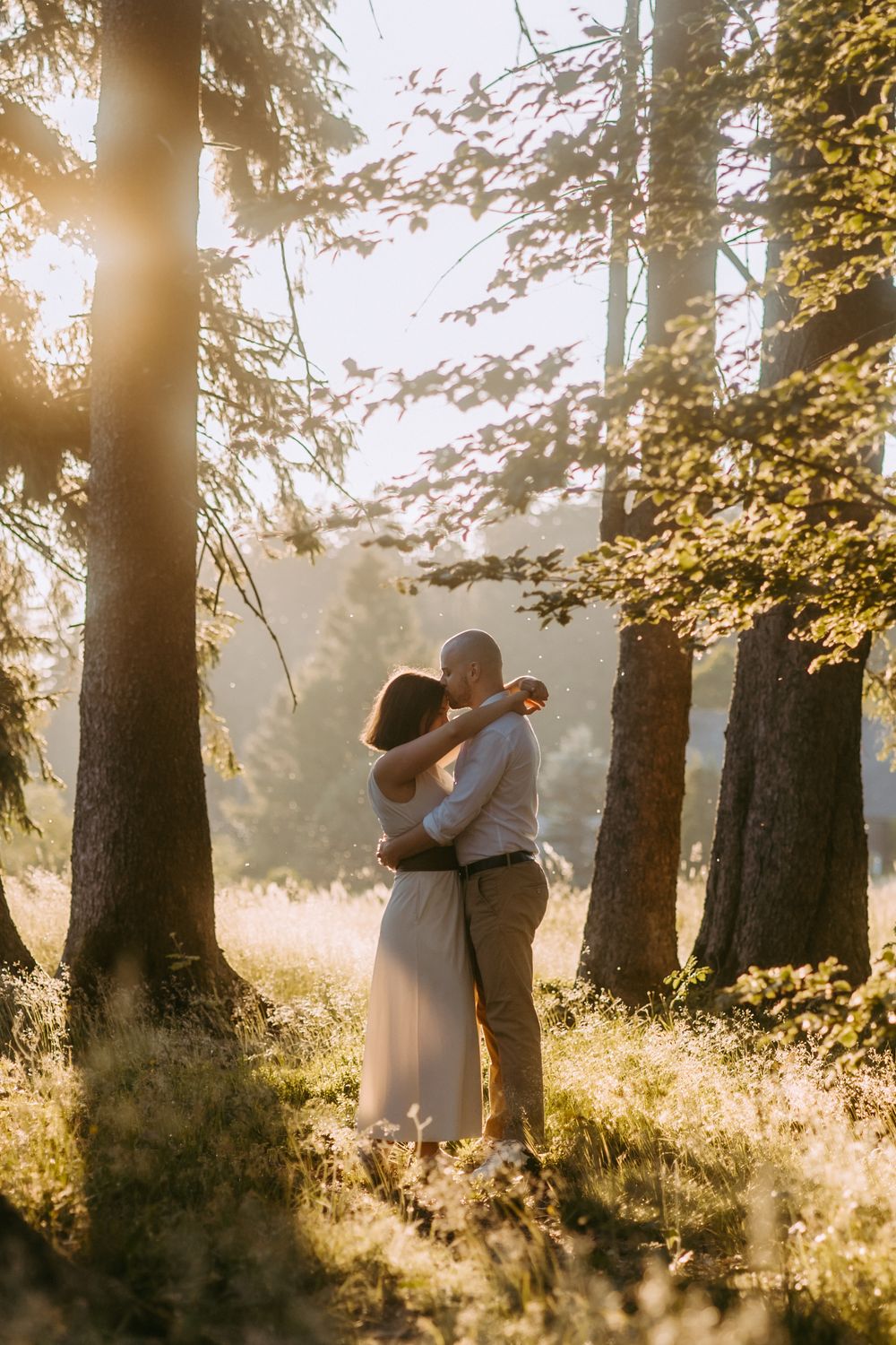 Couple embracing in sunlit forest with golden light filtering through tall trees.