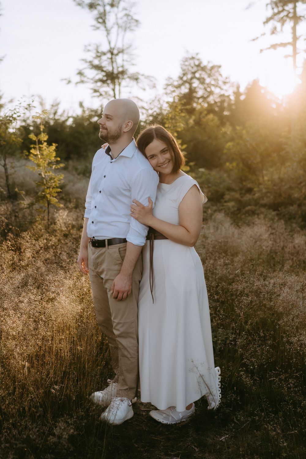 Couple embracing in golden sunset light among tall grass and trees, woman in white dress smiling at camera.